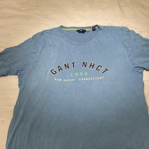 Blå t-shirt från Gant - Säljer en blå t-shirt från Gant med tryck 'GANT NHCT 1949 New Haven Connecticut' på framsidan. T-shirten har en rund halsringning med en grön detalj på insidan av kragen. 