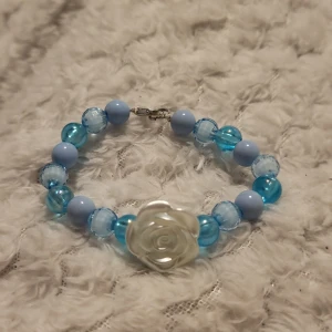 Blått armband med rosdetalj - Charmigt armband med blå pärlor i olika nyanser och en vit rosdetalj i mitten. Perfekt för att ge en färgklick till din outfit.