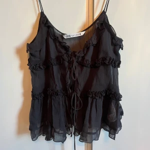 Svart volanglinne från Zara - Snyggt svart linne från Zara med volangdetaljer och tunna axelband. Toppen har en luftig och lätt design med knytning framtill, snyggt för en stilren look. Perfekt att bära en varm sommar dag!