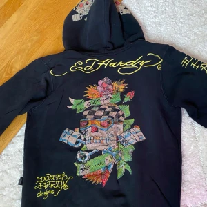 Svart hoodie från Ed Hardy - Säljer en svart hoodie från Ed Hardy med färgglada broderade detaljer och en stor dödskalle på ryggen. Hoodien har dragkedja framtill och långa ärmar. Använd men fortfarande i bra skick. Några stenar har ramlat av men det är inget som syns. Passar storlek S eller liten M. Är nog lite oversize på storlek XS.