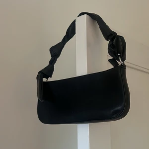 Svart axelväska med knytband - Snygg svart axelväska i skinn med unika knytband på axelremmen. Perfekt för att ge en stilren touch till din outfit. Väskan har en minimalistisk design och är lätt att matcha med olika stilar.