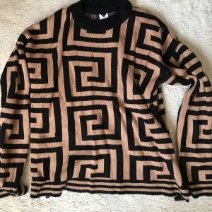 Svart och beige mönstrad tröja från Visual Clothing Project - Säljer en unik svart och beige tröja från Visual Clothing Project med ett geometriskt mönster. Tröjan har långa ärmar och en rund halsringning. Perfekt för att sticka ut i mängden!