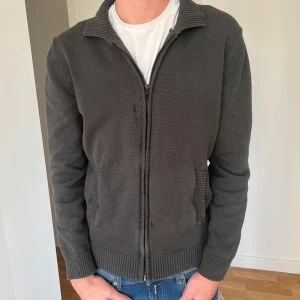 Stickad zip up från dressman - Säljer en stilren grå stickad jacka med dragkedja och ribbade detaljer. Perfekt för kyligare dagar med sin långärmade design och bekväma passform.