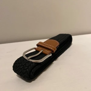 Svart flätat bälte (grisch bälte) - Snyggt svart flätat bälte med bruna läderdetaljer och en stilren silverfärgad spänne. Också kallat Grisch bälte. Perfekt för att ge en extra touch till din outfit. Passar både till jeans och chinos. Passar både tjejer och killar i alla åldrar då de är väldigt Stretchigt och töjbart. 