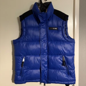 Blå dunväst från Plein Sport - Säljer en blå dunväst från Plein Sport med svart axelparti och dragkedja. Västen har ett stort silverfärgat tigertryck på ryggen och en camouflagemönstrad insida. Sparsamt använd. 