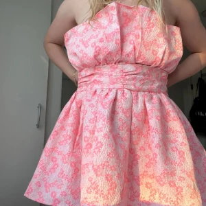 Rosa axelbandslös klänning - Säljer en söt rosa axelbandslös klänning med blommigt mönster. Klänningen har en markerad midja som ger en fin siluett. Perfekt för sommaren. Storlek 40 men skulle mer säga 38. Aldrig använd