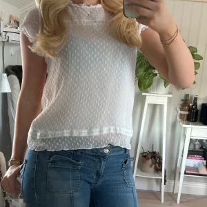 Vit blus med spetsdetaljer - Säljer en söt vit blus från Only med spetsdetaljer och korta ärmar. Blusen har ett diskret prickigt mönster och en fin volangkant vid ärmslut och nederkant. Perfekt för en somrig look!