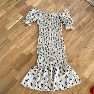 Blommig klänning med volang - Charmig blommig klänning med volangdetaljer och puffiga kortärmar. Klänningen har en smickrande passform och är perfekt för somriga dagar. Den är i en ljus färg med svarta blommor som ger en romantisk touch.