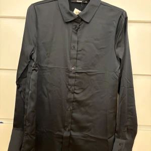 Svart långärmad skjorta - Snygg svart skjorta i mycket bra skick. Den har en klassisk krage och knappar längs framsidan. Perfekt för både vardag och festliga tillfällen. Skjortan är långärmad och har en stilren design som passar till det mesta. Aldrig använd, lappen kvar