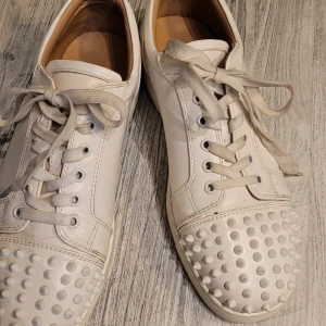 Vita sneakers med nitar - Säljer ett par vita sneakers i skinn med coola nitar fram. De har snörning och en snygg röd sula.. Använda men i bra skick.