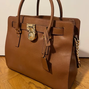 Brun handväska med gulddetaljer från Michael kors - Säljer en super snygg brun handväska från Michael Kors i mycket bra skick. Väskan har eleganta gulddetaljer och både ett kortaste och längre handtag. Den är lagom stor och har plats för allt du behöver🤎