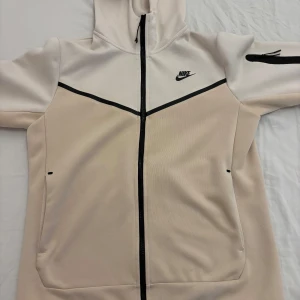 Beige och vit hoodie från Nike - Säljer en snygg beige och vit hoodie från Nike i mycket bra skick. Den har en dragkedja framtill och en huva för extra mys. Perfekt för höst och vår! Passar både till träning och chill. Det går att sänka priset.