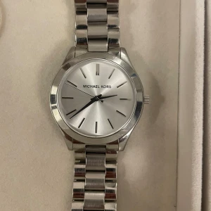 Silverfärgad klocka från Michael Kors - Snygg och stilren klocka i silver från Michael Kors. Den har en klassisk design med en metallarmband och en enkel urtavla. Perfekt för både vardag och fest. Klockan är i rostfritt stål och har en säker låsning. Ett måste för den modeintresserade!