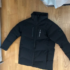 Svart dunjacka från Sail Racing - Säljer en svart dunjacka från Sail Racing i mycket bra skick. Jackan har en stilren design med dragkedja och logotypdetaljer på bröstet och ärmen. Perfekt för kalla vinterdagar med sin varma och bekväma passform. Passar både till vardags och mer äventyrliga aktiviteter.