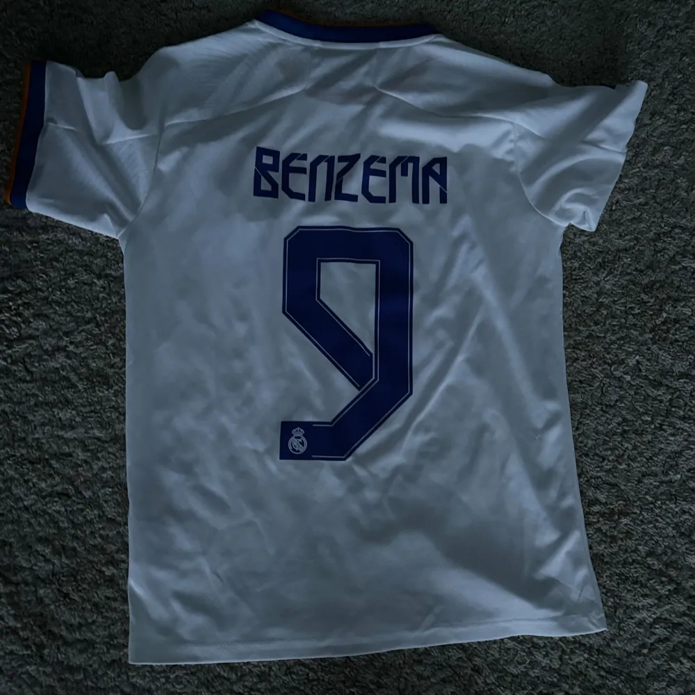 Säljer en vit Real Madrid fotbollströja från Adidas med korta ärmar. Den har blå detaljer och klubbens emblem på bröstet. På baksidan står det 'Benzema' och nummer 9. Perfekt för fans som vill visa sitt stöd! ⚽. T-shirts.