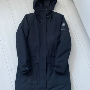 Mörkblå  parkajacka från Peak Performance - Säljer en svart parkajacka från Peak Performance i mycket bra skick. Jackan har en huva och långa ärmar, perfekt för kyliga dagar. Den är stilren och funktionell med diskreta detaljer. Passar både till vardags och mer aktiva dagar. Perfekt för höst och vinter!