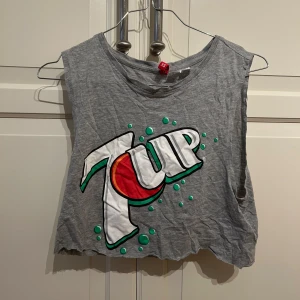Grå 7UP croppad linne - Säljer en cool grå croppad linne med stort 7UP-tryck på framsidan. Perfekt för sommarens varma dagar eller en avslappnad look. Linnet är ärmlöst och har en rå kant nertill för en edgy stil. Passar perfekt till jeans eller shorts!