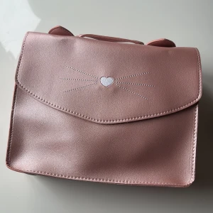 Rosa ryggsäck med kattdetalj - Söt rosa ryggsäck  från Yuko B med ett hjärta och kattmönster på framsidan. Väskan har en enkel design med en knappstängning. För barn 💖