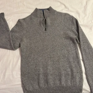 Grå cashmere half zip - Säljer en stilren grå ulltröja från Turo med dragkedja vid halsen. Perfekt för kyliga dagar med sin mjuka och varma känsla. Den har en snygg kontrast med blått på insidan av kragen. Passar både till vardags och lite finare tillfällen. Tröjan är Storlek S men passar xs