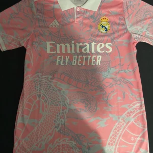 Rosa Real Madrid fotbollströja  - Säljer en snygg rosa Real Madrid fotbollströja från Adidas i storlek S. Tröjan har ett coolt drakmönster och korta ärmar. Tröjan har aldrig varit använd och är i bra material