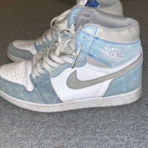 Nike Air Jordan 1 i blått och vitt - Säljer ett par Nike Air Jordan 1 i blått och vitt. Skorna har en snygg kombination av mocka och skinn med klassisk snörning. Perfekta för både vardag och fest. De är i bra skick med lite slitage på sulan.
