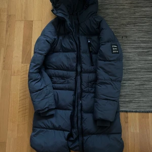 Mörkblå dunjacka med huva - Säljer en mörkblå lång dunjacka från Zink, perfekt för kalla vinterdagar. Jackan har en praktisk huva och dragkedja framtill. Den är i mycket bra skick och har en stilren design med en liten logga på ärmen. Perfekt för både vardag och vinteraktiviteter!