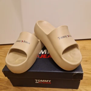 Beige sandaler från Tommy Jeans - Säljer ett par snygga beige sandaler från Tommy Jeans i storlek 38. De har en chunky flatform design som ger extra höjd och stil. Perfekta för sommardagar vid poolen eller stranden. Märket är tryckt på ovansidan för en trendig look. 