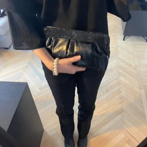 Svart kuvertväska med paljetter - Snygg svart kuvertväska i skinnimitation  med glittriga paljettdetaljer på kanten🙌 Perfekt för en utekväll eller fest! Väskans axelrem är enkel att dölja i väskan så du kan bära den som en clutch eller över axeln. Mycket bra skick! Se sista bild för längd på mig som är 163cm🌸