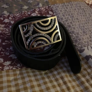 Svart bälte med glittrigt spänne - Säljer ett svart bälte med ett stort, glittrigt spänne i silverfärg. Bältet har en unik design med cirkulära mönster på spännet. Det finns lite slitage på utsidan av bältet, men det är fortfarande i bra skick som inte syns när man har på sig bältet 