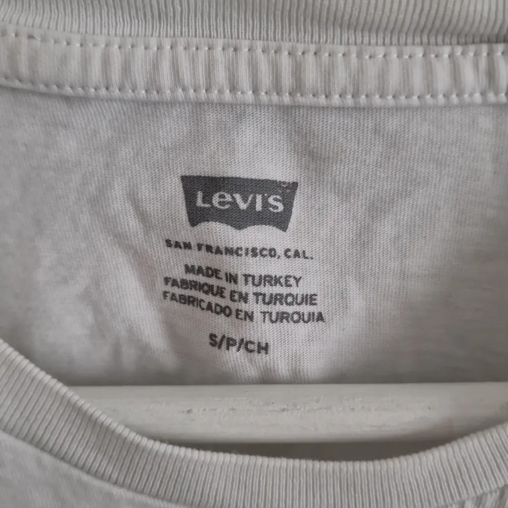 Säljer en klassisk vit t-shirt från Levi's i bomull. Den har en röd logga på framsidan och i bra skick (förutom knappt synliga fläckar, se bild) Perfekt för en avslappnad stil eller som basplagg i garderoben. Kortärmad och normal passform.. T-shirts.