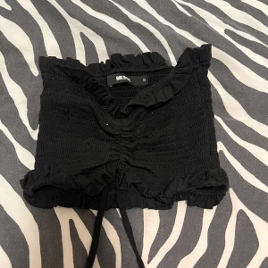 Svart smockad topp från Bik Bok - Säljer en supersöt svart smockad topp från Bik Bok i storlek S. Den har volangdetaljer och en knytning framtill som ger en fin passform. Perfekt för sommarens fester eller en dag på stan. Toppen är i mycket bra skick och redo för nya äventyr!