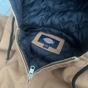 Brun jacka från Dickies - Säljer en snygg brun workjacka från Dickies i storlek L. Jackan har en dragkedja framtill och en huva med dragsko. Den är quiltad på insidan för extra värme och har ribbade muddar vid ärmslut och nederkant. Perfekt för höst och vår!