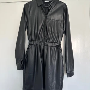 Svart klänning i skinnimitation från Noisy May - Säljer en supercool svart klänning i skinnimitation från Noisy May. Den har en elastisk midja och långa ärmar, perfekt för en edgy look. Klänningen har knappar framtill och en bröstficka som ger den en avslappnad stil. Perfekt för höst och vinter!
