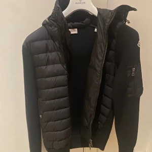 Moncler Wool blend down jacket - Svart Moncler jacka med huva, dun & stickade ärmar. Har dragkedja framtill och två fickor på sidorna. Jackan har även en ficka på vänster ärm. Mycket gott skick. Beställd från LuisaViaRoma för 8000kr. Inga skador eller defekter. Herr-modell