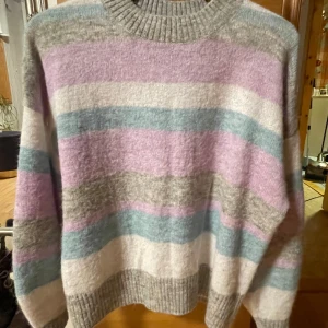 Randig stickad tröja i pastellfärger - Säljer en supermysig stickad tröja i pastellfärger. Den har breda ränder i lila, blått, beige och vitt. Perfekt för kyliga dagar med sin mjuka och varma känsla. Tröjan har en ribbad krage och långa ärmar. Passar perfekt till både jeans och kjol för en avslappnad look!