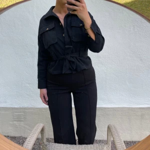 Svart jacka med knytband - Säljer en stilren svart shirt jacka med knytband i midjan. Jackan har långa ärmar och praktiska fickor med knappar framtill. Liten i storleken och aldrig använd. Perfekt för både vardag och fest!