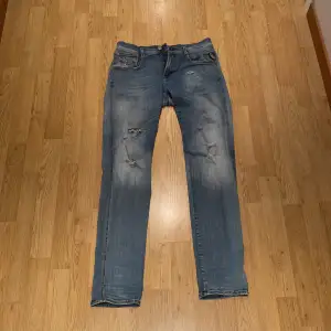 Säljer ett par snygga blå jeans med coola slitningar på knäna och låren. De har en klassisk femficksdesign och en knappgylf. Perfekta för en avslappnad stil!