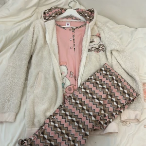 Mysig rosa pyjamas med björnmotiv - Supermysig pyjamas i rosa med ett gulligt björnmotiv. Setet inkluderar en långärmad tröja och byxor med ett zigzag-mönster i rosa och brunt. Tröjan har knappar framtill och en söt björn på bröstet. Perfekt för kalla nätter!