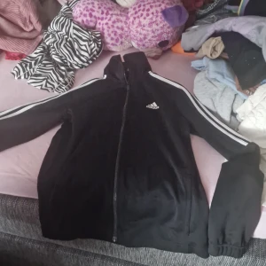 Svart träningsjacka från Adidas - Säljer en svart träningsjacka från Adidas i storlek S. Jackan har en dragkedja framtill och långa ärmar med vita ränder längs sidorna. Perfekt för träning eller som en casual vardagsjacka. Den är i bra skick och redo för nya äventyr!