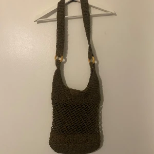 Brun stickad axelväska - Säljer en superfin brun stickad axelväska med ett unikt nätmönster. Väskan har en cool design med trädetaljer på axelremmen och är perfekt för en bohemisk look. Den är rymlig och har ett foder inuti för extra hållbarhet. Perfekt för sommarens äventyr eller en dag på stan!