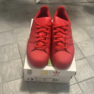 Röda Adidas Superstar Supercolor J - Säljer ett par snygga röda Adidas Superstar Supercolor J designade av Pharrell Williams. Skorna är i nyskick och har en klassisk snörning. Perfekta för att ge din outfit en färgklick!