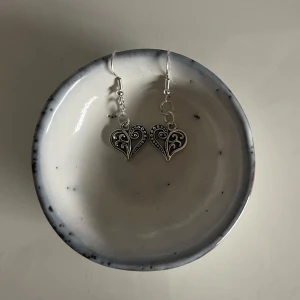 Silverfärgade hjärtformade örhängen - Säljer ett par superfina silverfärgade örhängen i form av hjärtan. De har ett detaljerat mönster med små prickar och snirkliga detaljer som ger dem en unik look. Perfekta för att piffa upp vilken outfit som helst! 