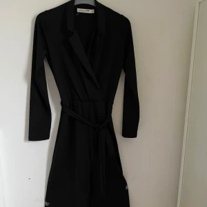 Svart långärmad klänning från Junkyard - Säljer en elegant svart långärmad byxsdress från Junkyard. Den har en stilren design med ett knytband i midjan för en snygg passform. Perfekt för både vardag och fest!