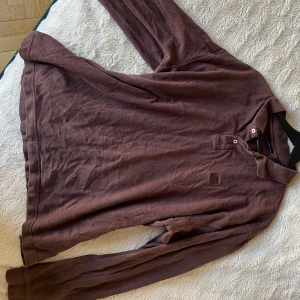 Brun pikétröja från FILA - Säljer en mysig brun pikétröja från FILA i storlek XXL. Tröjan har långa ärmar och en klassisk krage med knappar. Perfekt för en avslappnad och snygg höst stil! Den är i bra/worn skick och redo för en ny ägare!