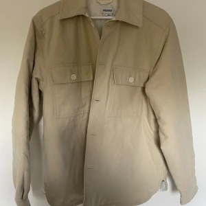 Beige overshirt från Weekday - Säljer en snygg beige overshirt från Weekday. Den har en quiltad insida för extra värme och stil. Två stora bröstfickor med knappar och en klassisk krage. Perfekt för lager-på-lager under hösten eller våren. Passar dig som är runt 185cm.