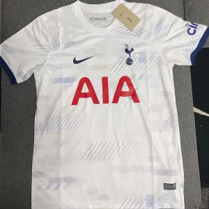 Vit Tottenham Hotspur fotbollströja - Säljer en vit Tottenham Hotspur fotbollströja från Nike. Tröjan har korta ärmar och är dekorerad med subtila grå mönster. Den har klubbens emblem och Nike-loggan på bröstet samt AIA som sponsor i rött. Perfekt för alla Spurs-fans!