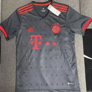 Säljer en snygg grå fotbollströja från Adidas med röda detaljer. Tröjan har korta ärmar och ett diskret mönster över hela tyget. Perfekt för fotbollsfans som vill ha något stilrent och sportigt. Stor logga på bröstet och tre ränder på axlarna. Passar perfekt till match eller träning!