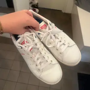 Säljer ett par klassiska vita Adidas Stan Smith sneakers med röda detaljer. Skorna har snörning och är i skinn. Perfekta för både vardag och fest! De är i bra skick och redo för en ny ägare. Storlek 38 och 2/3. 