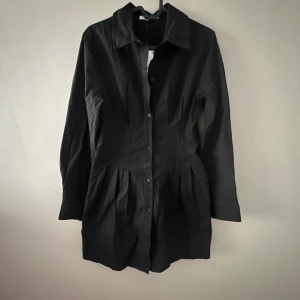 Svart klänning från Pull&Bear - Snygg svart klänning från Pull&Bear i storlek S. Den har en klassisk krage och knappar framtill. Perfekt för både vardag och festliga tillfällen. Klänningen är i nyskick och har en stilren design som passar till allt. Den är figur sydd.