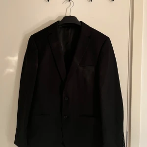 Svart kavaj herr - Snygg svart kavaj, perfekt för både bal och fest. Kavajen har en klassisk design med två knappar framtill och en stilren ficka på bröstet. Insidan är elegant fodrad med röd detalj.